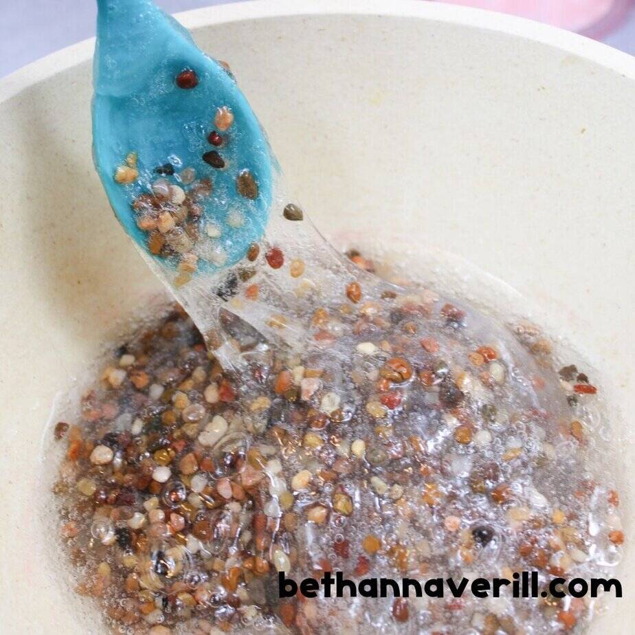 A hand holding a small clear bowl of contact solution over a mixing bowl filled with clear slime and colorful pebbles, with a teal spoon in the bowl.