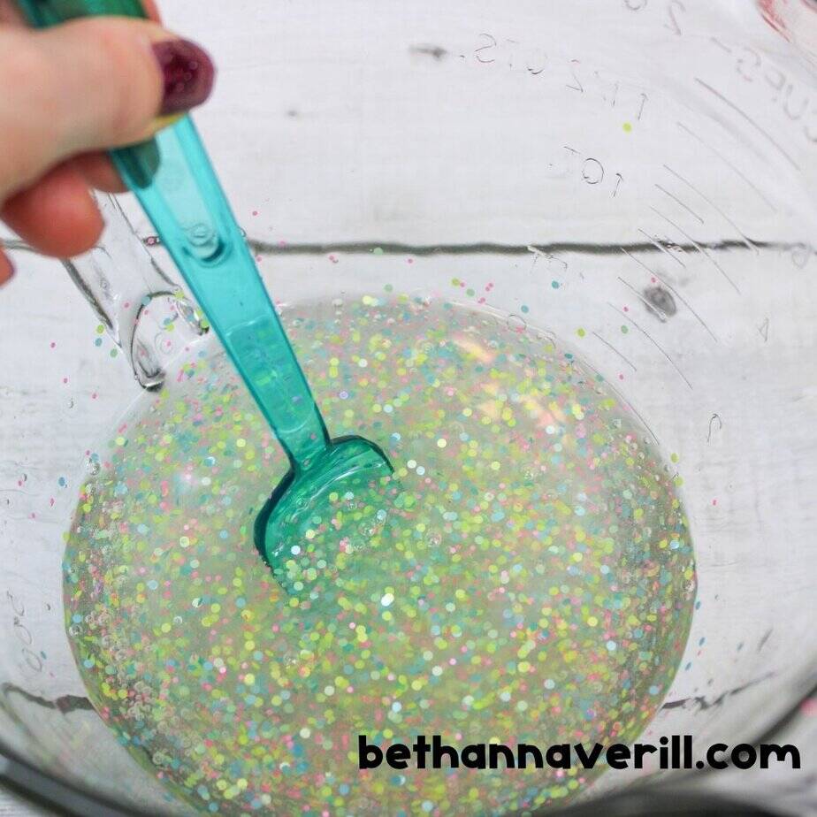 A teal spoon stirs pastel-colored glitter into clear slime inside a large glass mixing bowl, blending the sparkles throughout.
