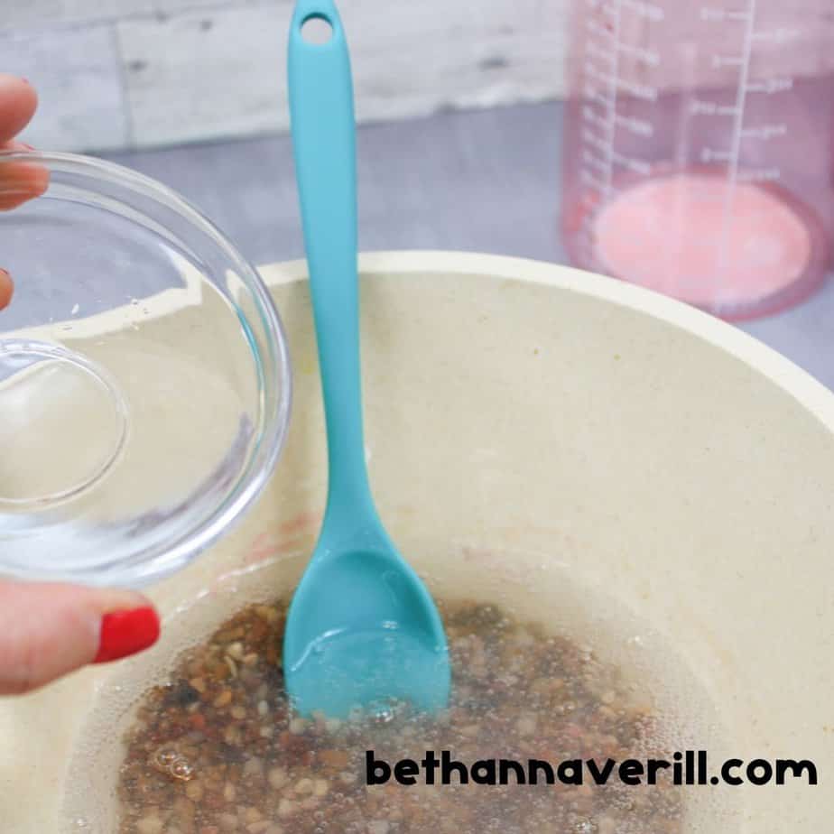A teal spoon lifting sticky, clear slime mixed with multicolored pebbles from a bowl, showing the gooey texture and embedded stones.