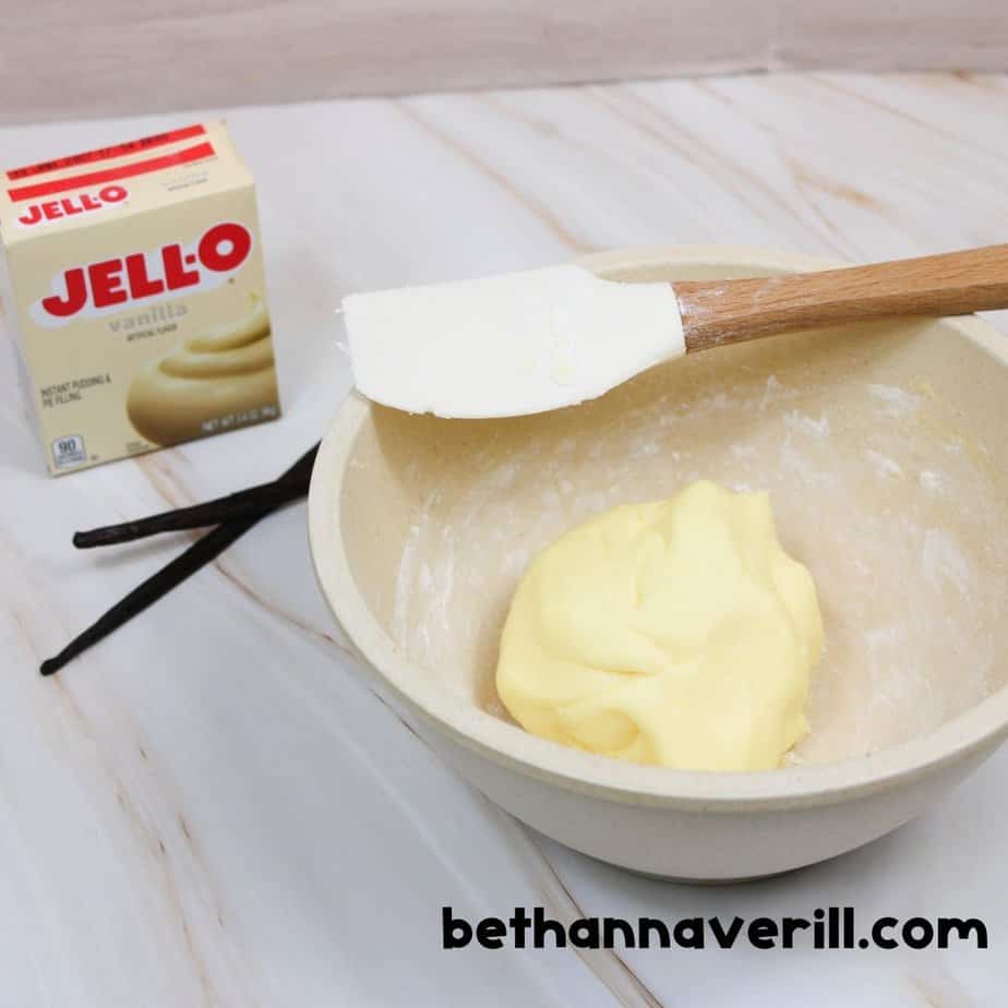 Beige mixing bowl with a ball of vanilla pudding slime inside, a spatula resting on the edge, and a box of Jello vanilla pudding with vanilla beans nearby. bethannaverill.com branding displayed.