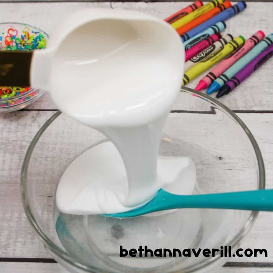 White glue being poured into a bowl to start making crayon slime, with a spoon and crayons visible nearby.