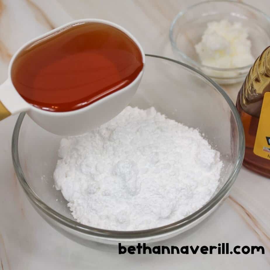 A full white measuring cup of golden honey being poured over powdered sugar in a glass bowl, with coconut oil and a honey bottle nearby. bethannaverill.com is shown at the bottom