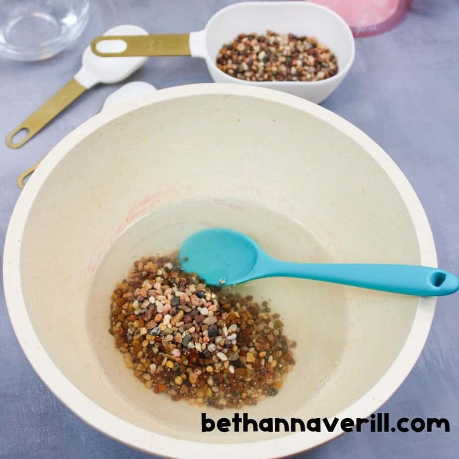 A cream-colored bowl with clear slime and a pile of small colorful pebbles just added, ready to be mixed with a teal spoon.
