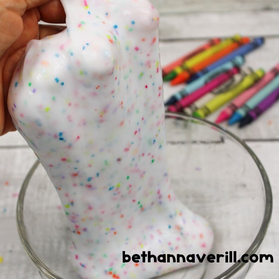 Stretchy white crayon slime with colorful flecks being lifted from a glass bowl, with crayons scattered in the background.