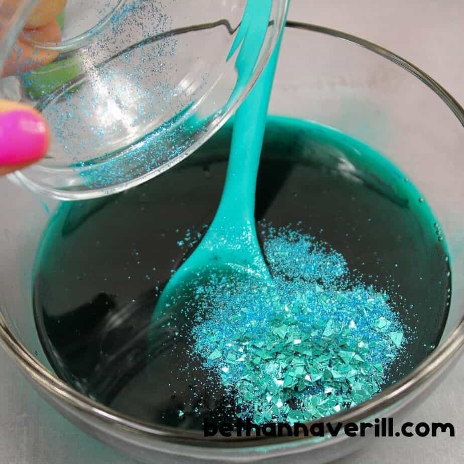 A bowl of teal slime is topped with a colorful mix of chunky mermaid-themed glitter, including pastel stars, tails, and iridescent shapes, being stirred with a turquoise spoon.