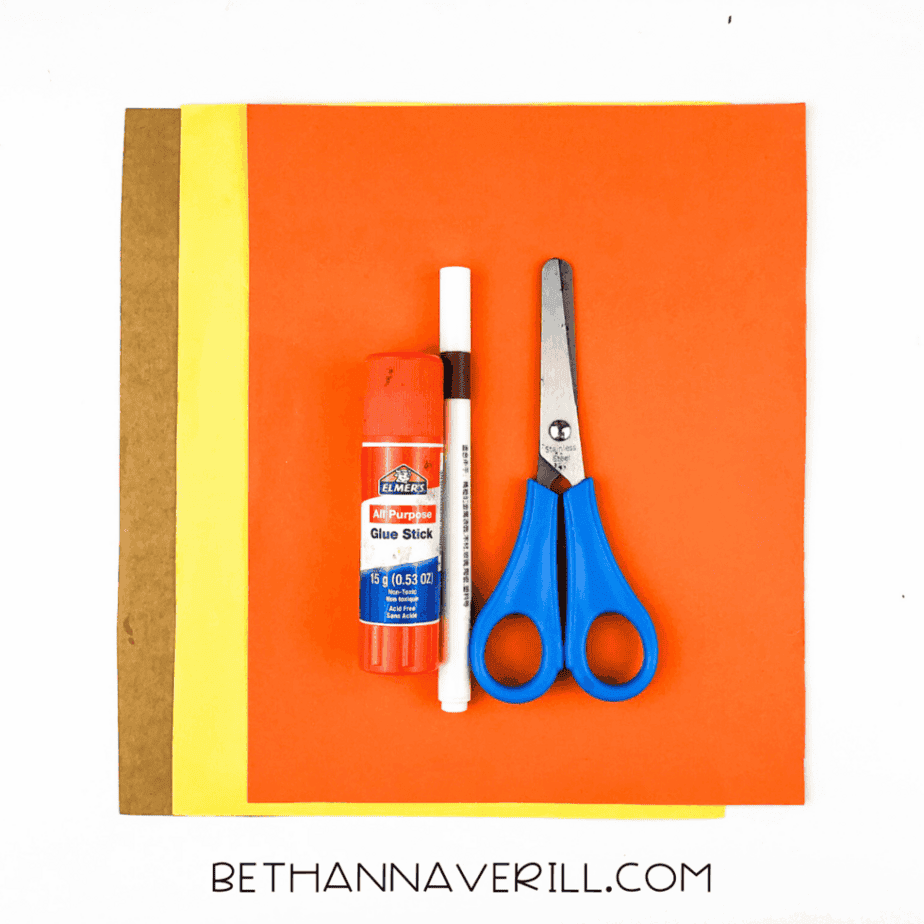 A flat lay of orange, yellow, and brown paper with glue stick, brown marker, and blue-handled scissors.