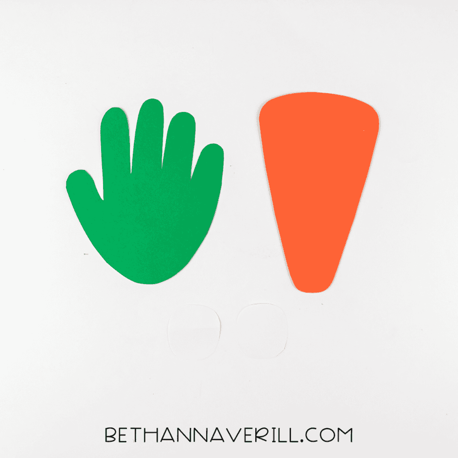 Green handprint cutout, orange carrot shape, and white eye pieces laid out