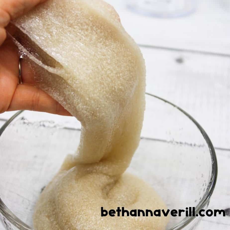 A hand stretches gritty sand slime upward, showing its soft stretch and grainy texture in a glass bowl.