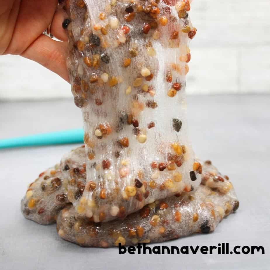 A hand stretching sticky pebble slime upward, showing its gooey, stretchy texture and the embedded multicolored pebbles.