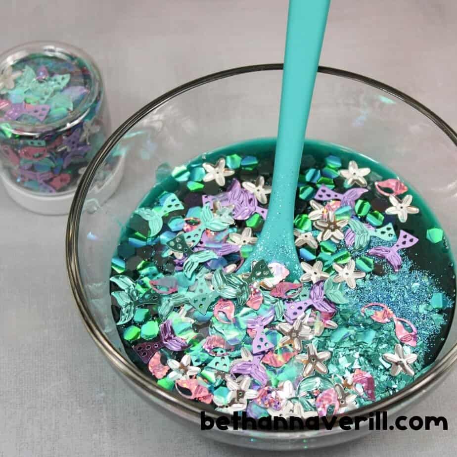 A bowl of teal slime is topped with a colorful mix of chunky mermaid-themed glitter, including pastel stars, tails, and iridescent shapes, being stirred with a turquoise spoon.