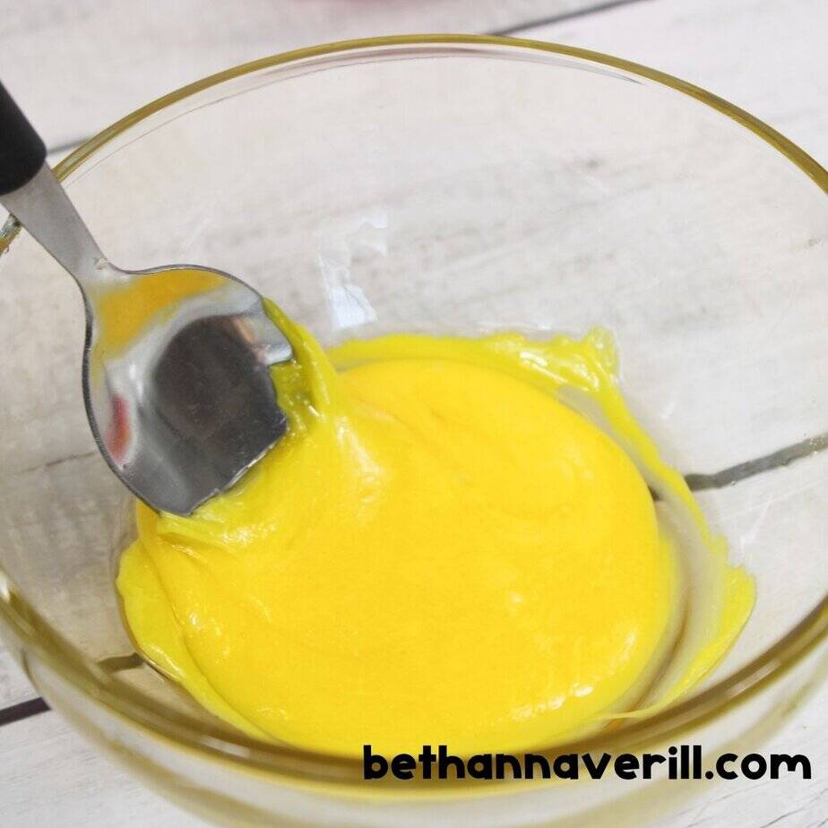Melted yellow Starburst being stirred with a spoon in a clear bowl after microwaving, showing gooey slime texture.