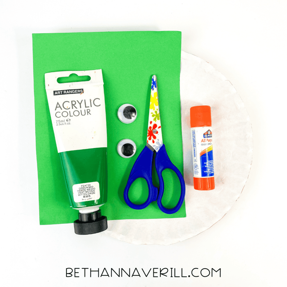 Supplies laid out for the alligator craft: green paint, scissors, googly eyes, glue stick, green paper, and a white paper plate.