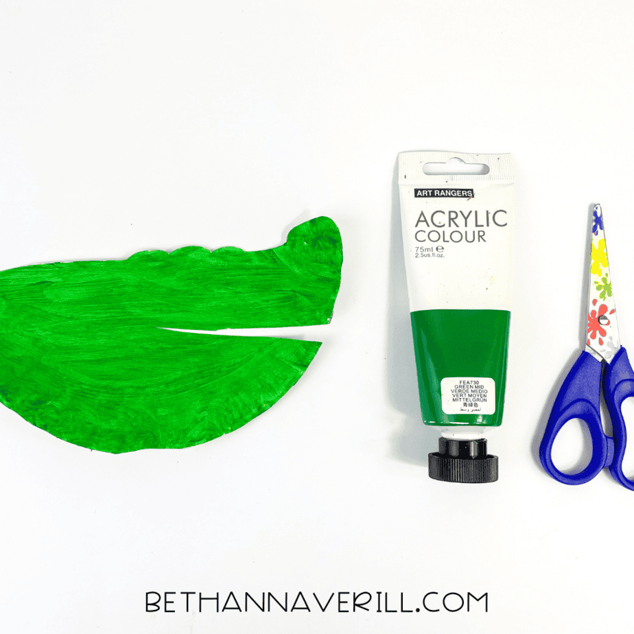 Paper plate painted bright green and cut into an alligator head shape, with paint and scissors beside it.