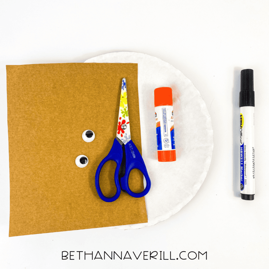 Materials for dung beetle craft including brown paper, googly eyes, scissors, glue, and a white paper plate.