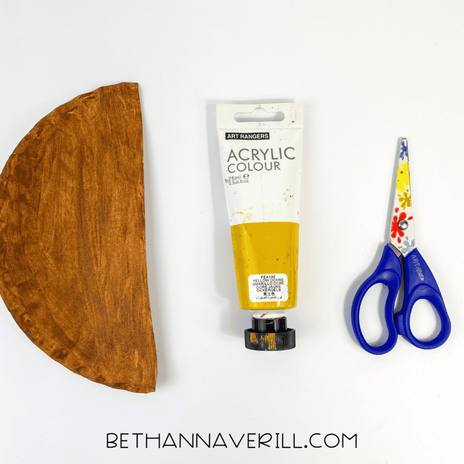 Half of a paper plate painted brown next to yellow paint and scissors.