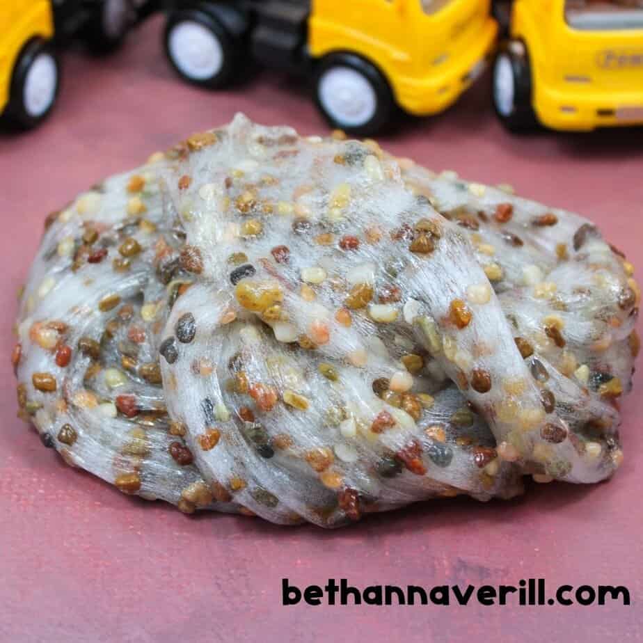 A close-up of clear slime mixed with small pebbles, swirled into a pile on a pink surface with yellow toy trucks in the background.