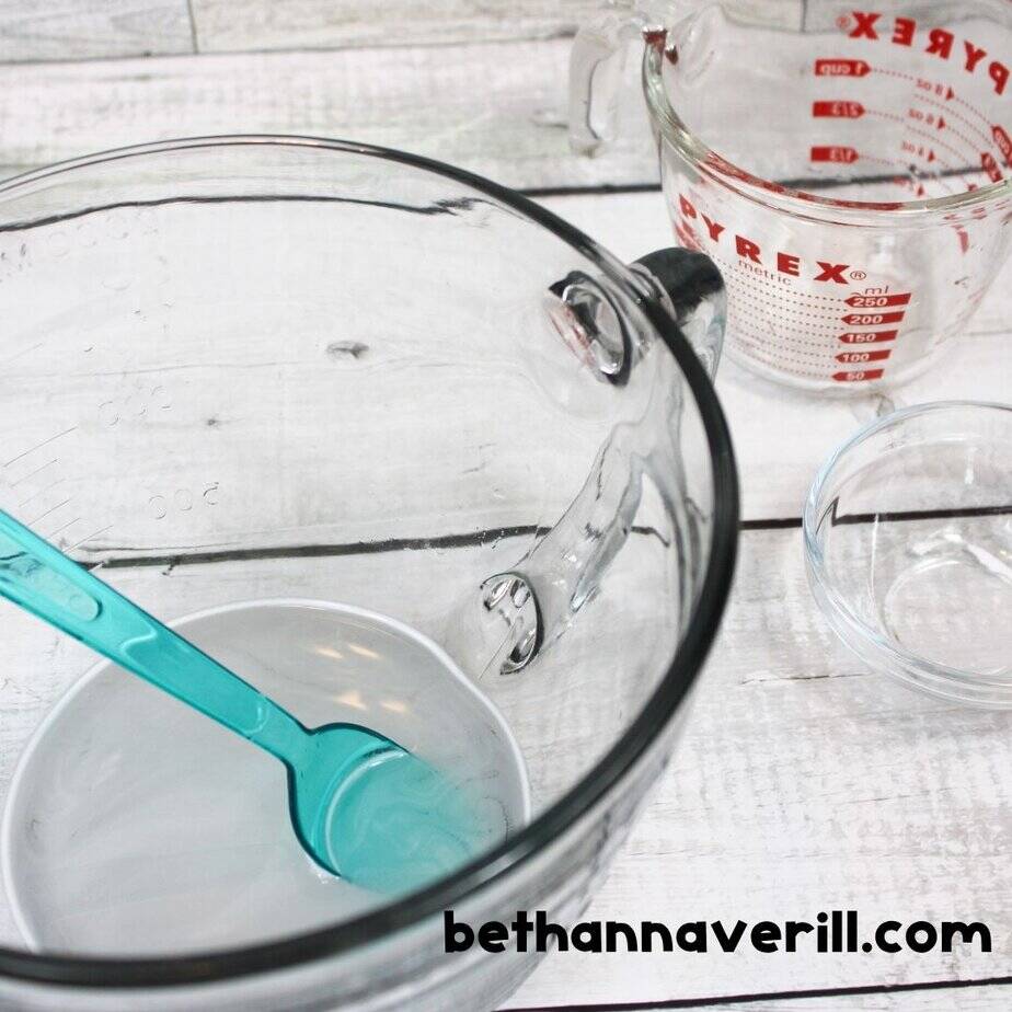 A glass bowl with water and baking soda being mixed using a teal spoon; additional bowls and measuring cups are nearby to make spring slime