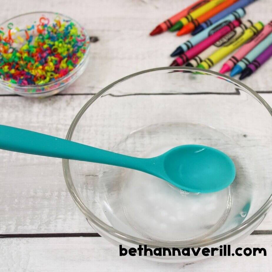 Glass bowl with spoon and water, next to a dish of crayon shavings and crayons, ready to mix in baking soda for slime.
