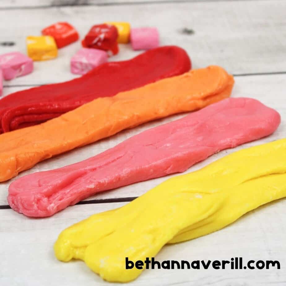 Four colorful strips of Starburst slime—red, orange, pink, and yellow—lined up on a white surface with unwrapped candies behind them.