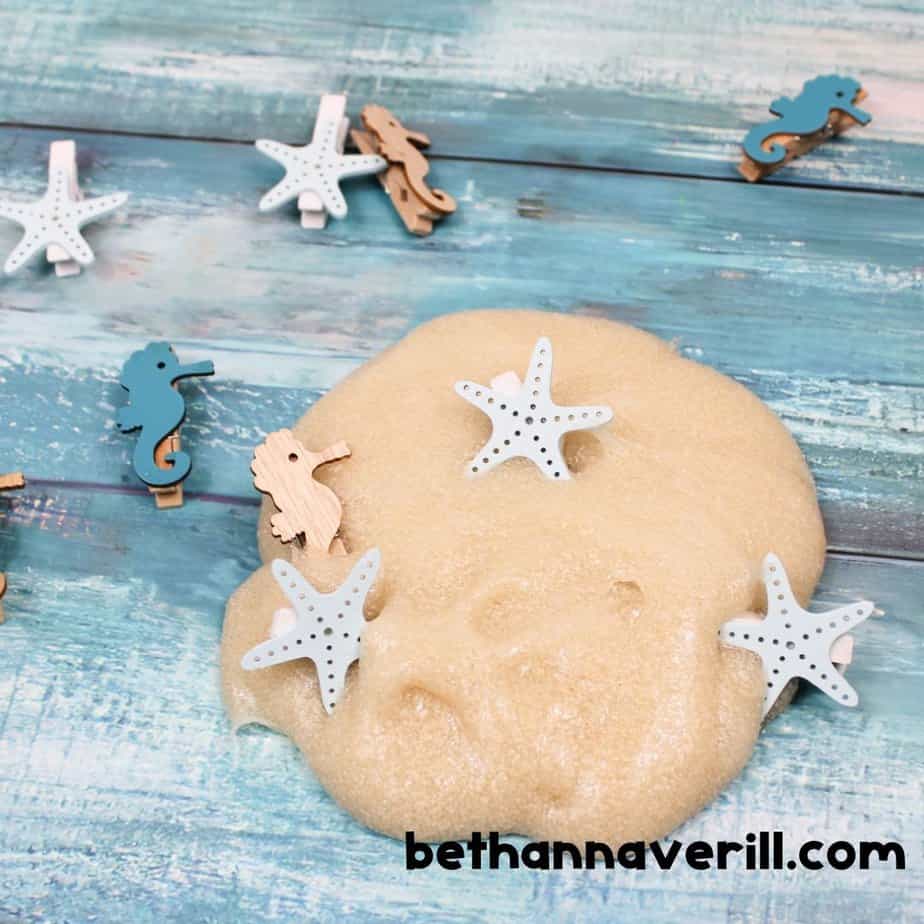 A tan blob of sand slime on a blue background, adorned with wooden starfish and seahorse clips, mimicking a mini beach scene.