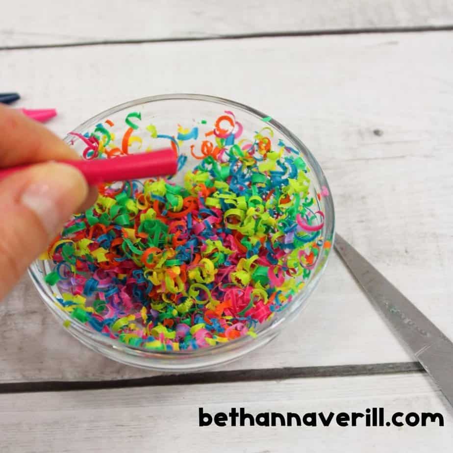 Hand holding a pink crayon over a bowl of colorful crayon shavings, showing the process of making crayon shavings for slime with scissors nearby.