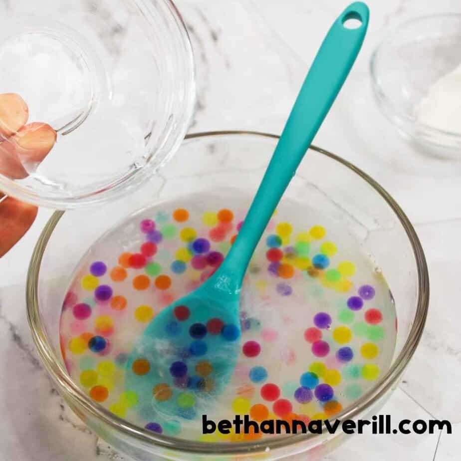 A hand is pouring clear liquid from a small bowl into a mixing bowl filled with water beads and slime solution, ready to activate the slime.