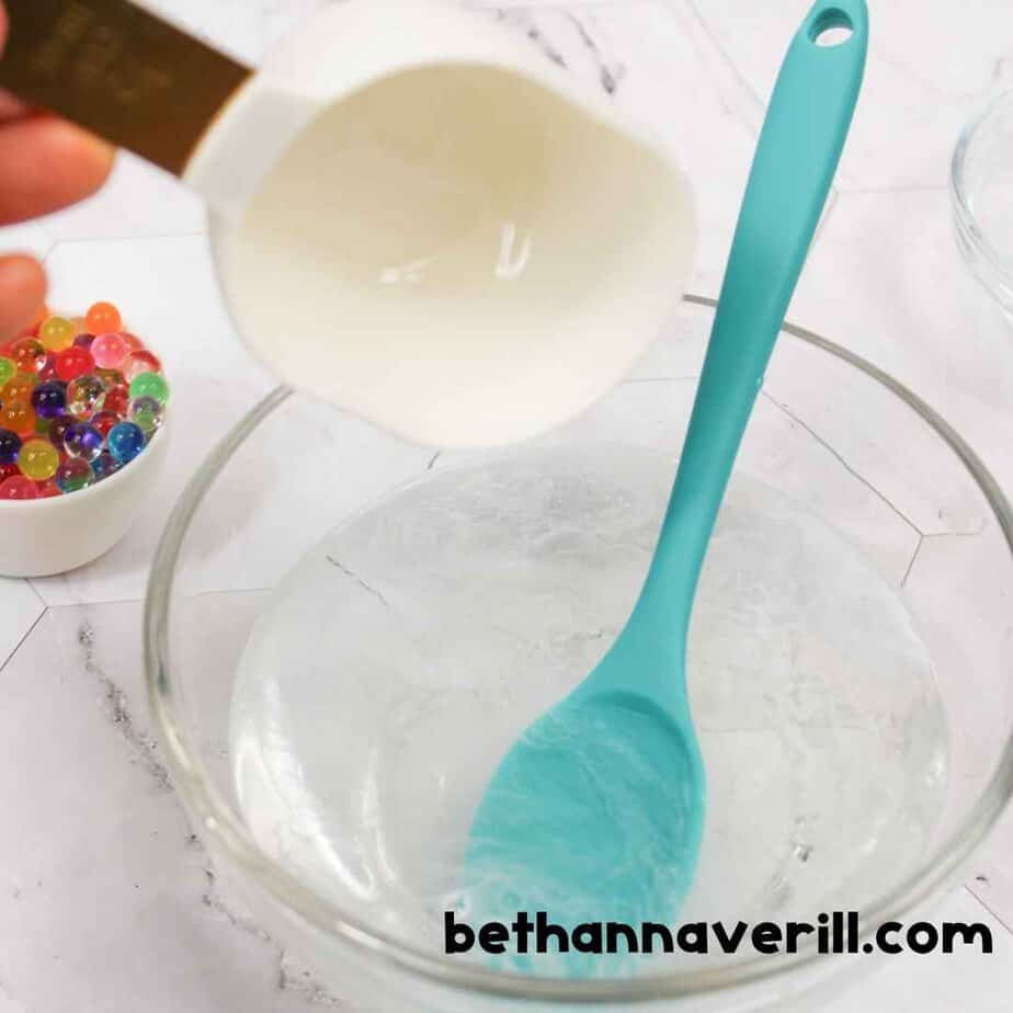 Clear glue is being poured from a white measuring cup into a glass bowl containing water, while a teal spoon is ready to stir. A small bowl of colorful Orbeez sits nearby.