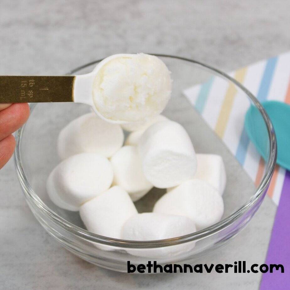 A tablespoon of coconut oil is held above a glass bowl filled with jumbo marshmallows, ready to be microwaved for edible slime making.