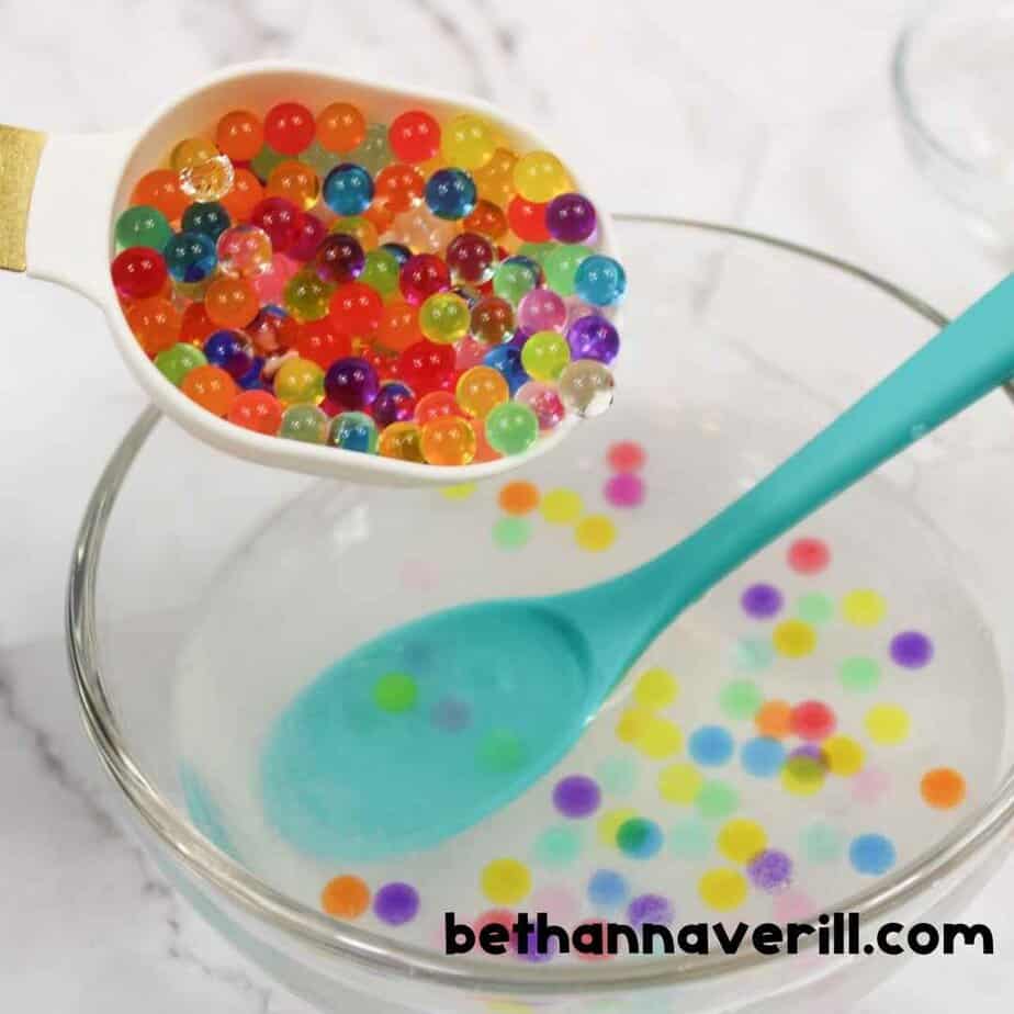A close-up of a spoon filled with vibrant water beads being added to a bowl of clear slime mixture, with a teal spoon already in the bowl.