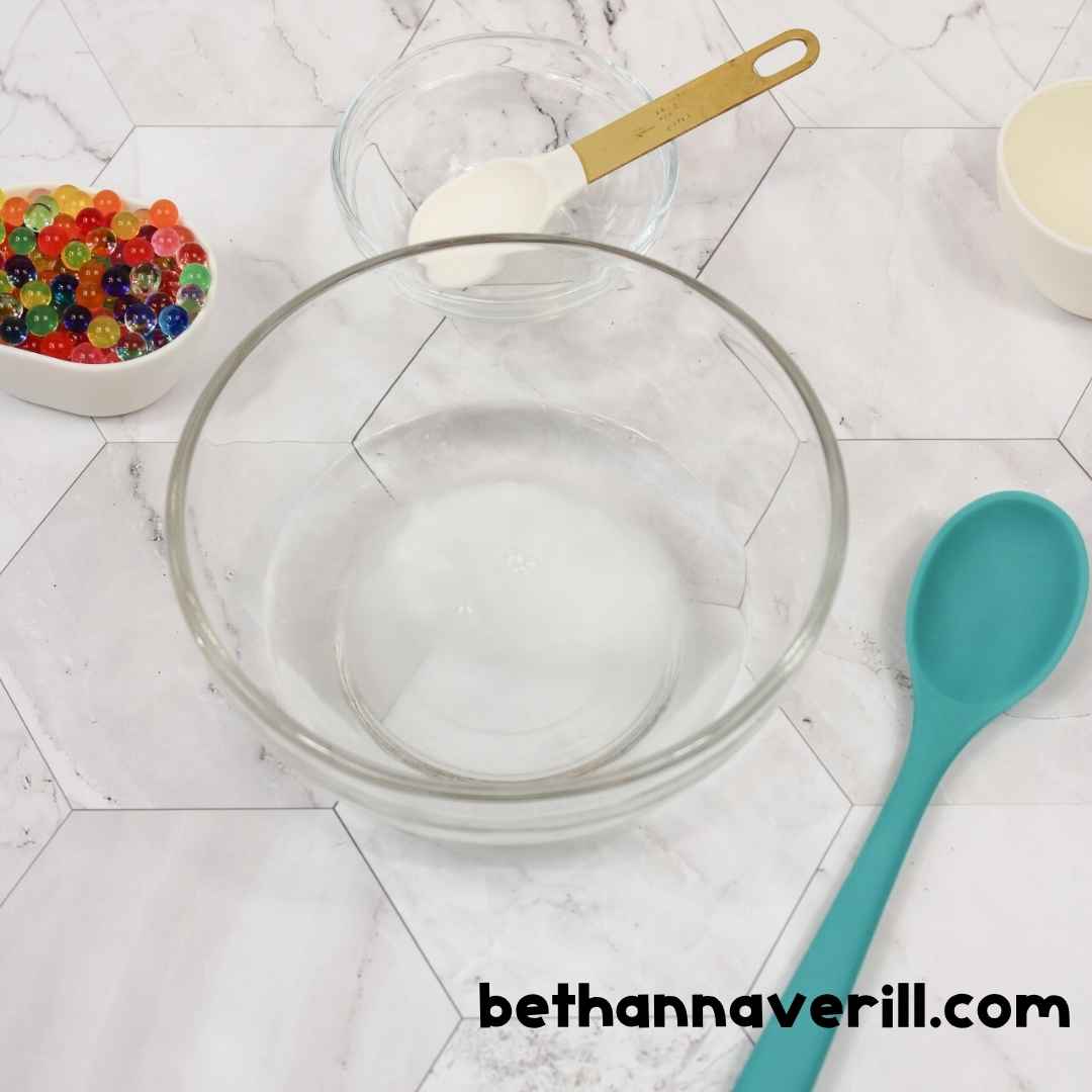 A clear mixing bowl filled with warm water is placed in the center, surrounded by small bowls of baking soda, glue, and water beads, along with a teal mixing spoon.