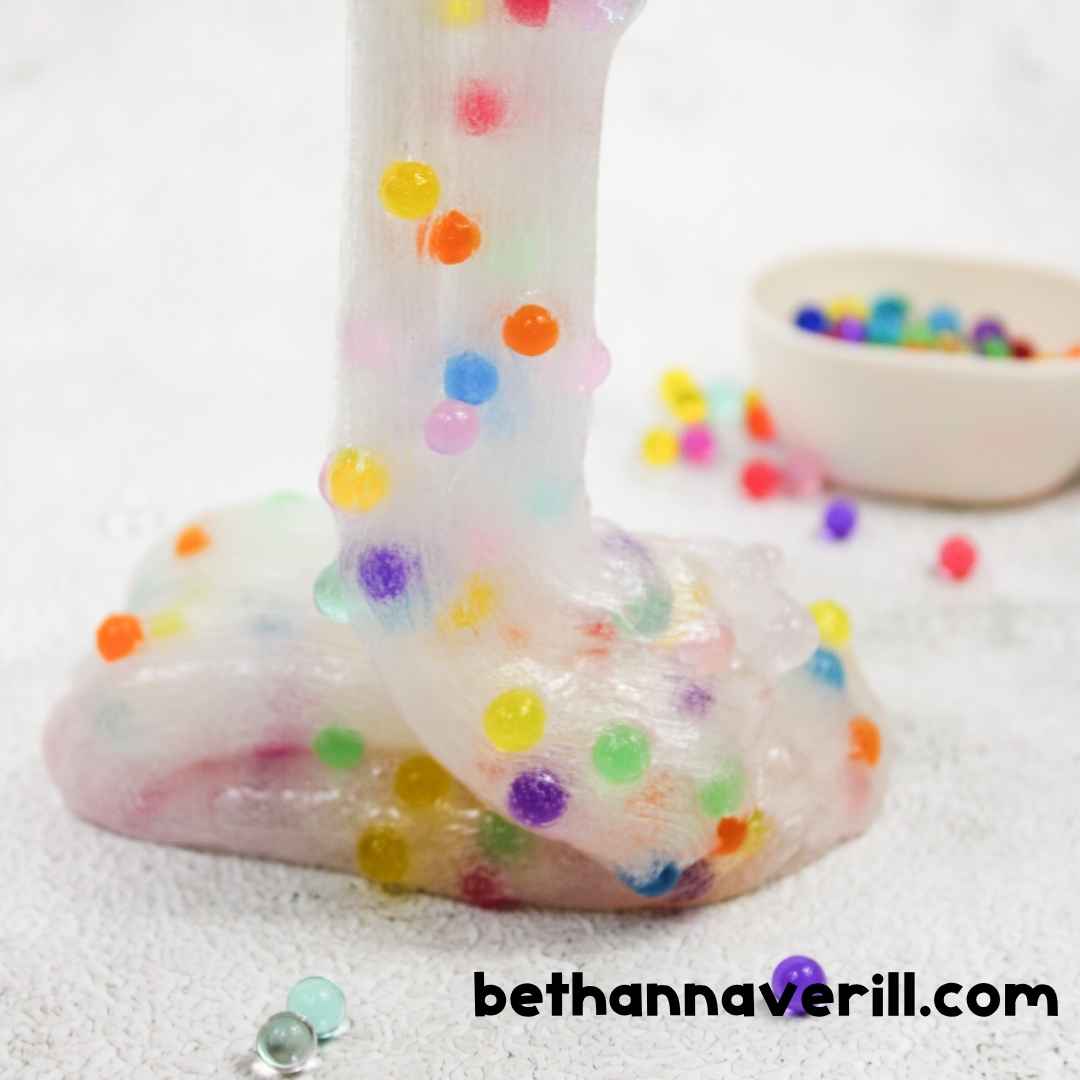 A hand stretches a long piece of glossy slime loaded with colorful water beads, showing the slime’s stretchy, gooey texture mid-pull.