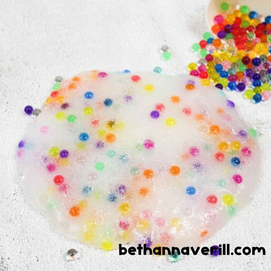 Clear slime spread on a white surface, filled with vibrant rainbow-colored water beads. A small bowl of loose beads is seen spilling nearby.