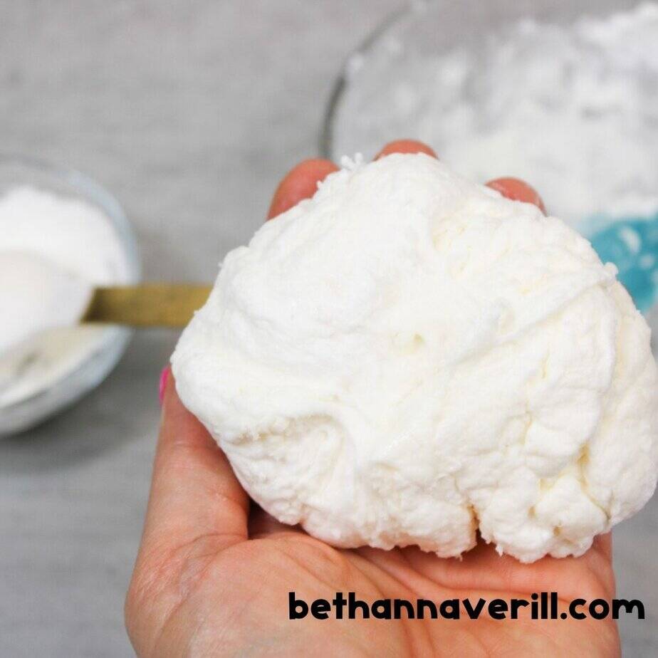 A hand holds a soft white clump of marshmallow slime, still a bit textured and stretchy, while cornstarch and a teal spoon sit in the background.