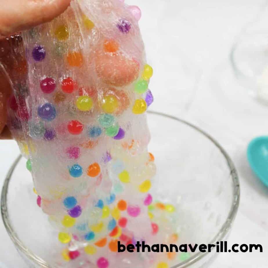 Close-up of a hand lifting stretchy clear slime filled with Orbeez-style water beads over a glass bowl, showing the sticky, bead-filled texture.