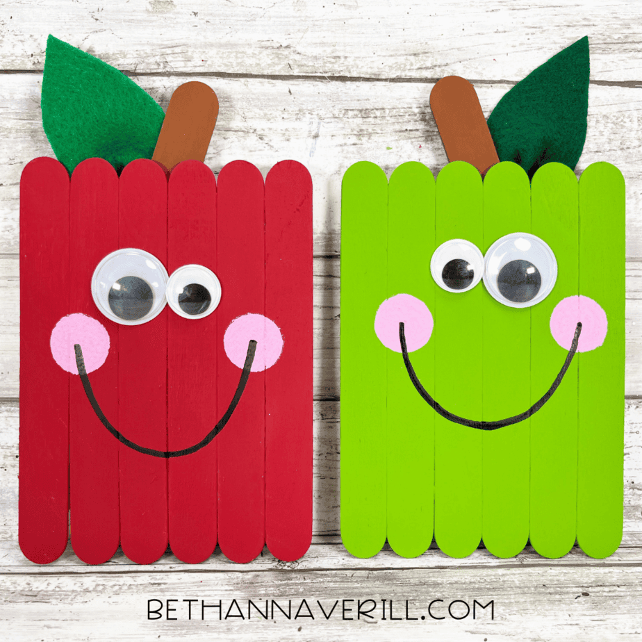 Finished red and green apple popsicle stick crafts with big googly eyes, pink cheeks, and smiling faces.