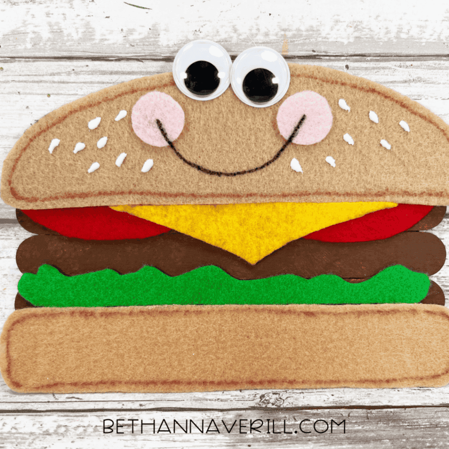 Finished cheeseburger popsicle stick craft with googly eyes, cheeks, smile, and sesame seed details.
