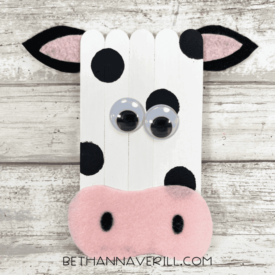 Completed popsicle stick cow craft with white body, black spots, pink snout, ears, and googly eyes.