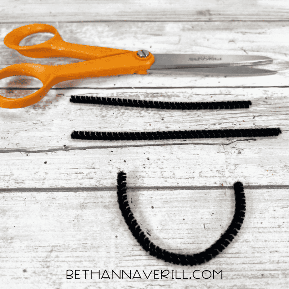A pair of scissors next to three pieces of cut black pipe cleaner, including one bent into a U-shape for a smile.