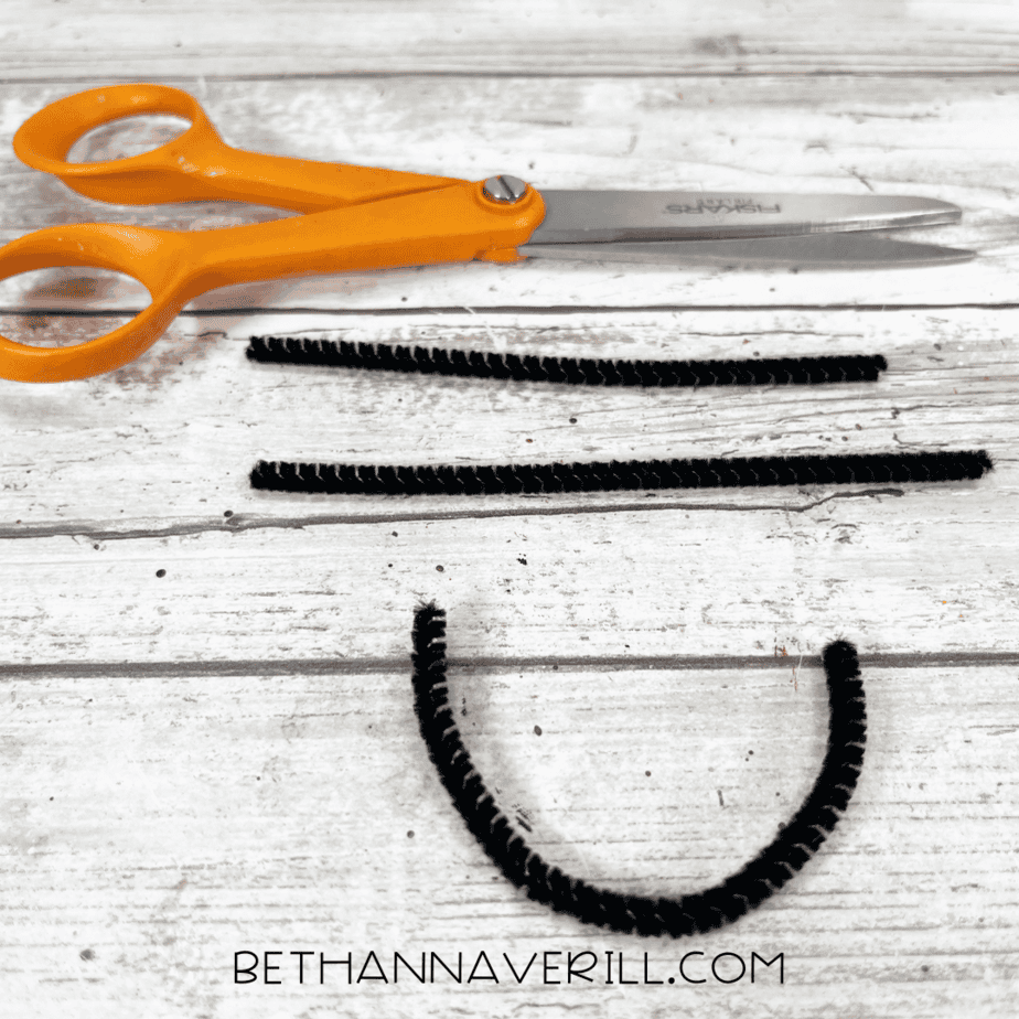 A pair of scissors next to three pieces of cut black pipe cleaner, including one bent into a U-shape for a smile.