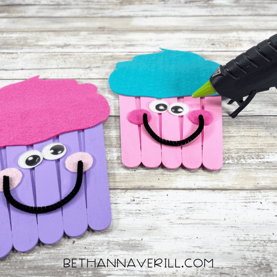Two finished cupcake crafts with googly eyes, felt cheeks, and smiling mouths made of pipe cleaners, being glued together.