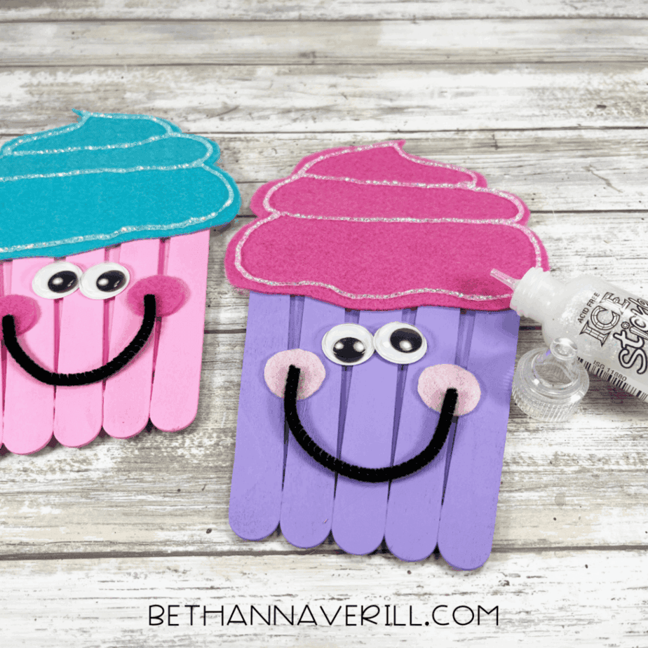 Two colorful cupcake crafts made from popsicle sticks, decorated with felt frosting and googly eyes, with a bottle of glitter glue nearby.