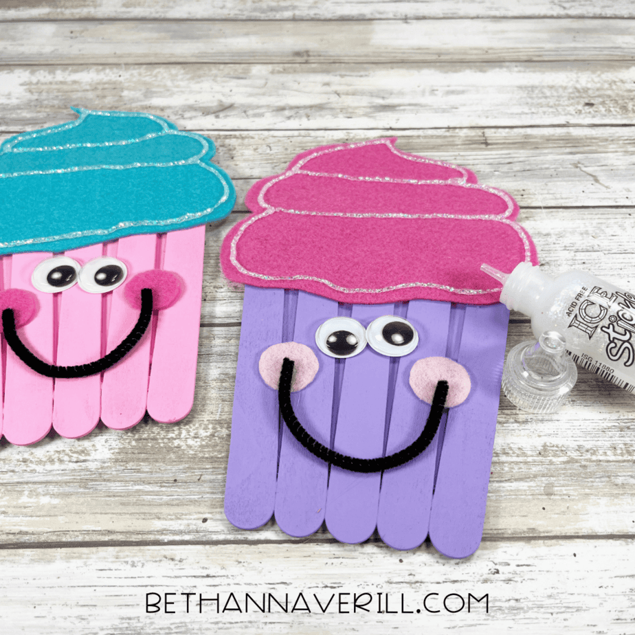 Two colorful cupcake crafts made from popsicle sticks, decorated with felt frosting and googly eyes, with a bottle of glitter glue nearby.
