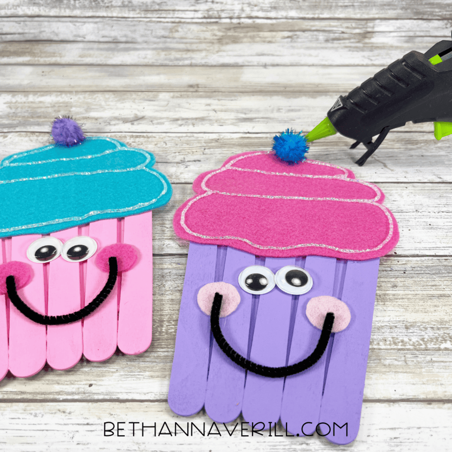 A hot glue gun attaching a blue pom-pom to the top of a purple and pink cupcake popsicle stick craft.