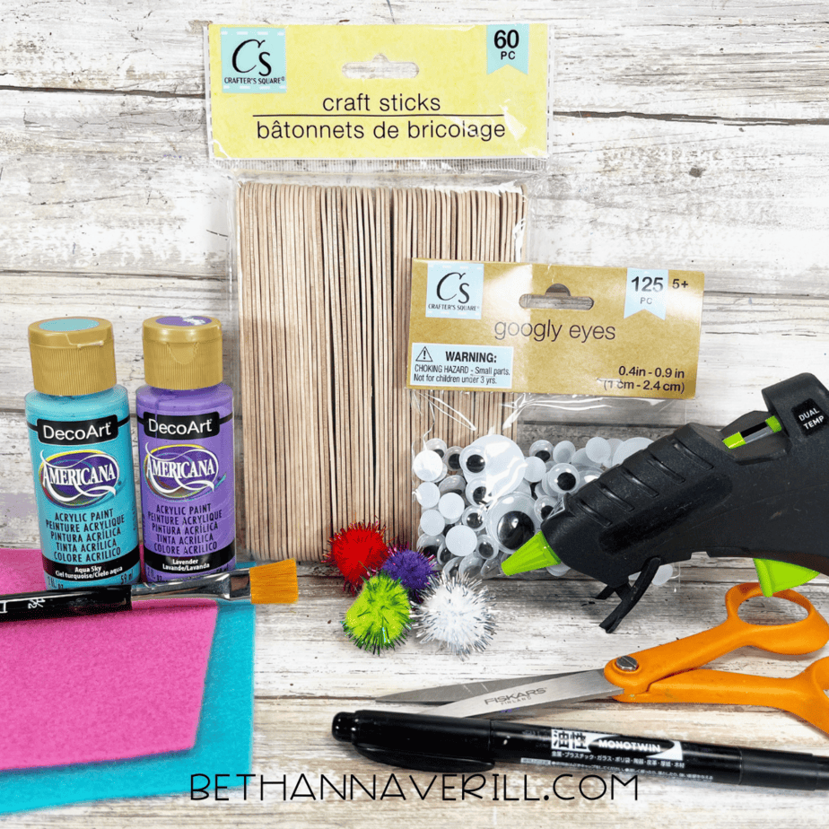 A variety of craft supplies laid out on a wooden surface, including paint bottles, googly eyes, pom-poms, felt sheets, scissors, a glue gun, craft sticks, and a marker.
