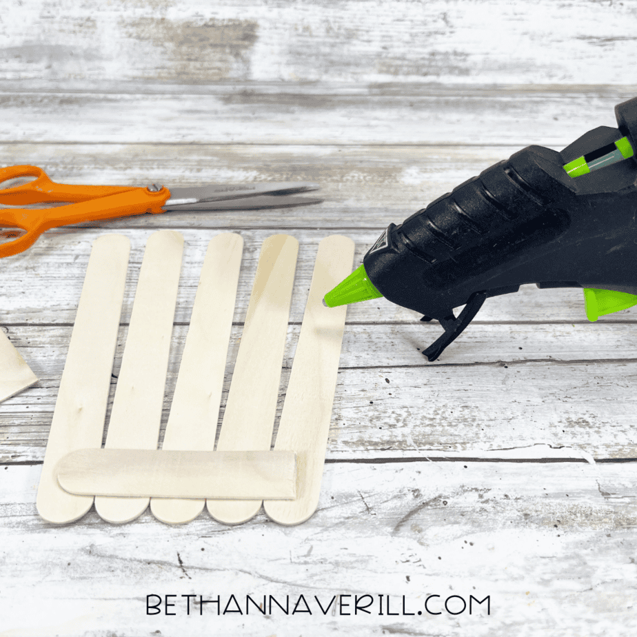Five wooden craft sticks lined up vertically and glued together at the bottom with a horizontal stick, next to a glue gun and scissors.