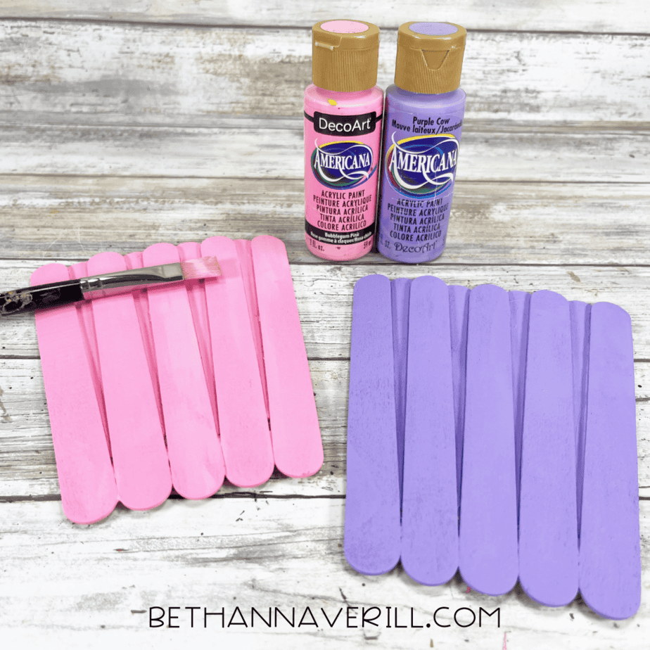 Two finished popsicle stick panels, one painted pink and one purple, shown next to their corresponding acrylic paint bottles and a paintbrush.