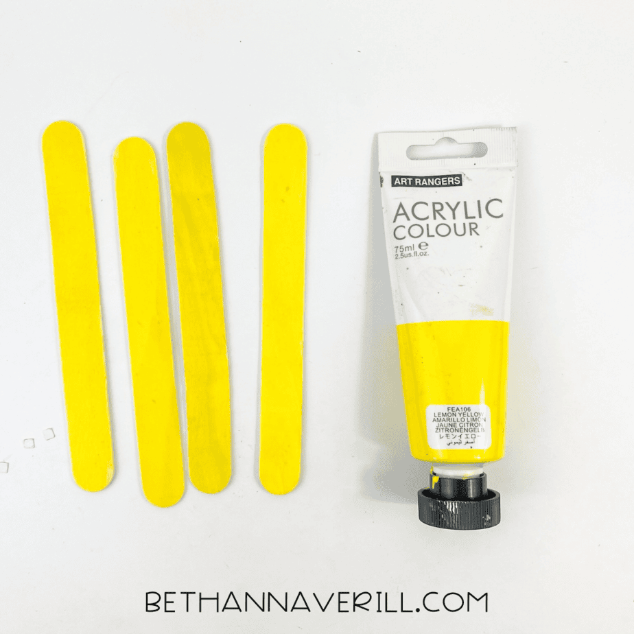 Four popsicle sticks painted yellow next to acrylic paint tube.