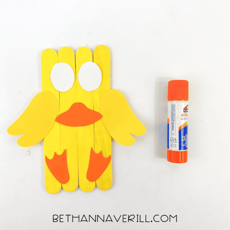 Duck craft in progress with wings, beak, feet, and white eyes glued onto yellow popsicle sticks.