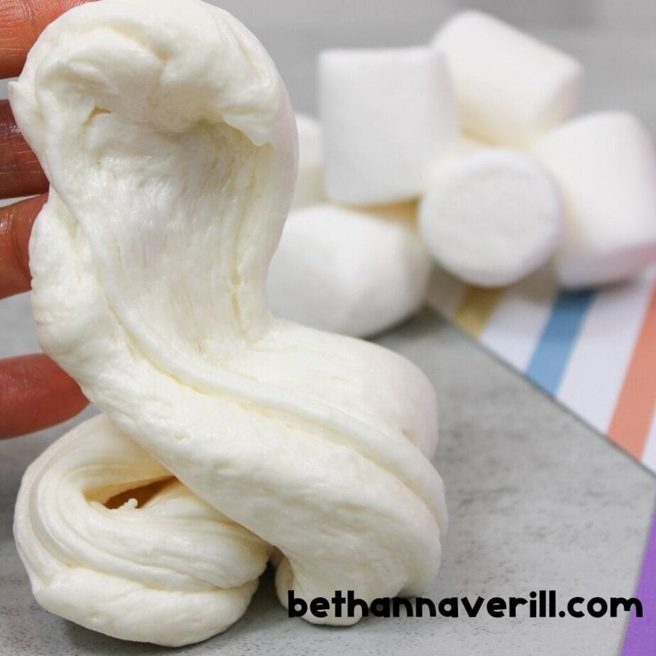 Thick edible slime made from marshmallows is stretched and folded, with jumbo marshmallows and colorful paper nearby on a gray countertop.