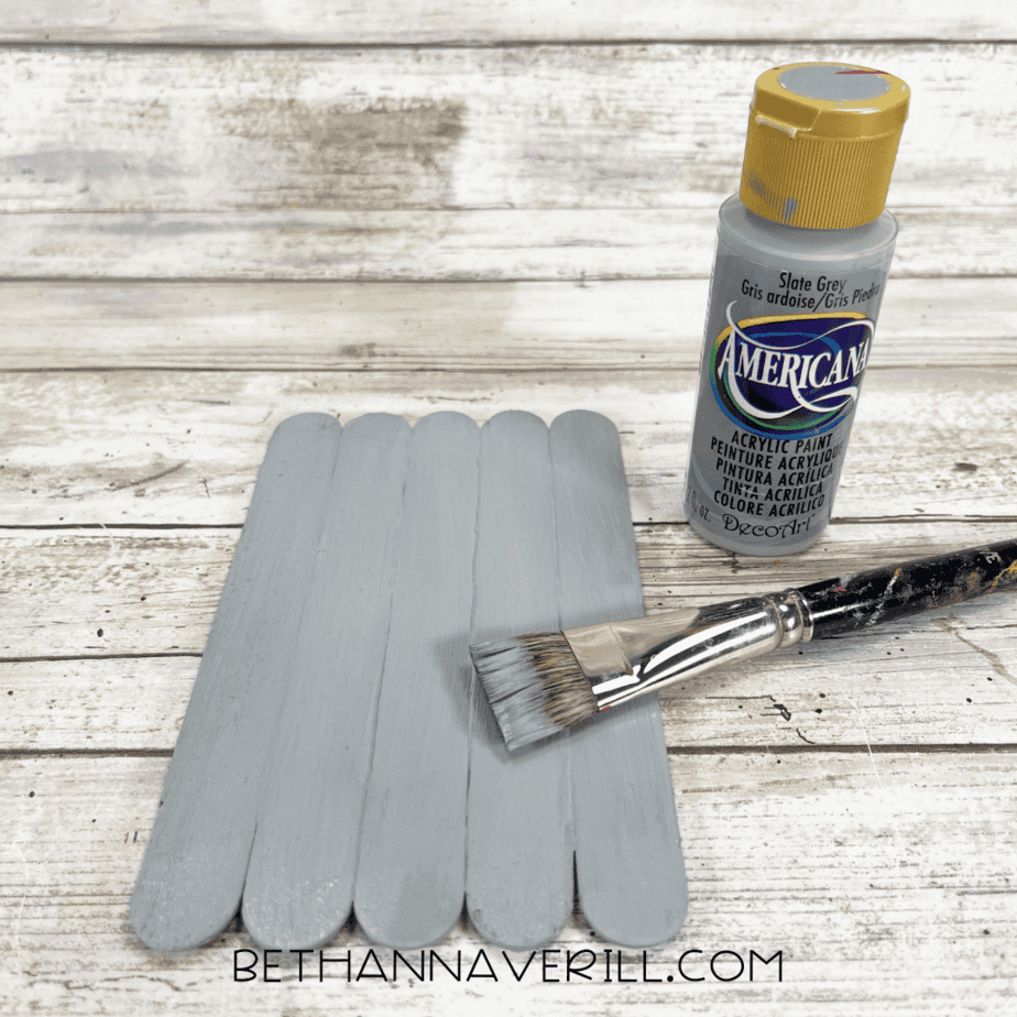 Grey painted popsicle sticks drying with paintbrush and paint bottle beside them.